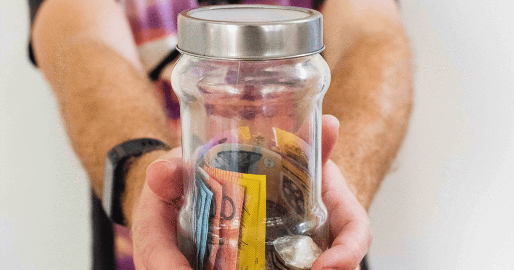 Closeup of a man's hands reaching toward the viewer holding a jar full of colourful money, bills and coins.