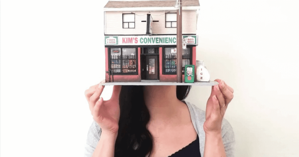 Image of a young woman holding up a model of the Kim's Convenience store in front of her face.