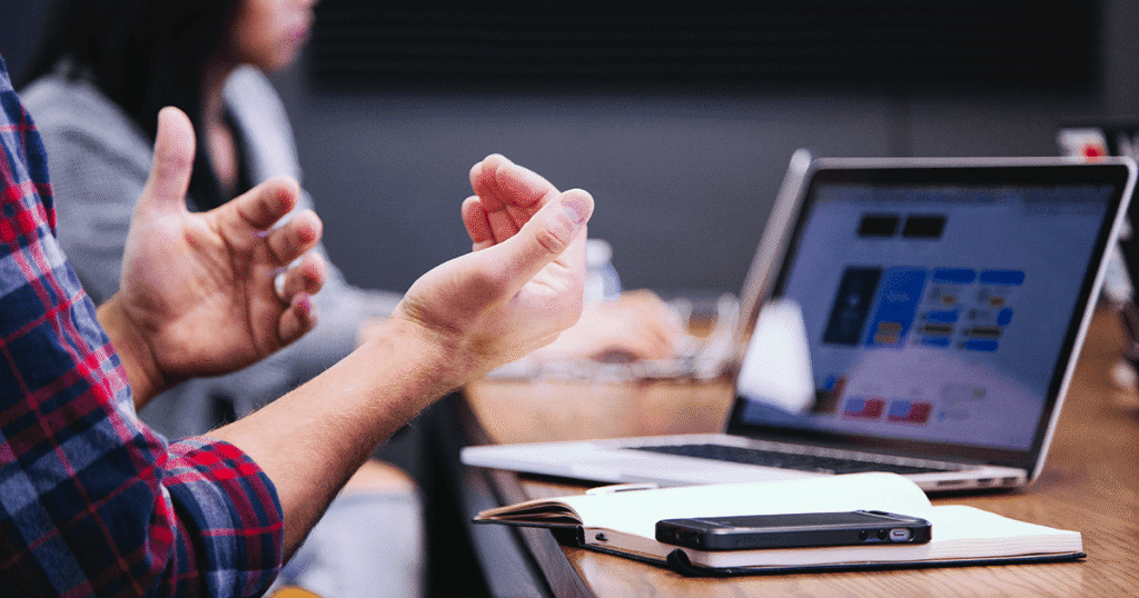Meeting room with laptops on table, a person's hands gesturing openly in discussion.