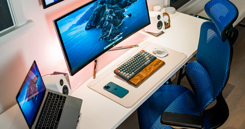 Overhead picture of a home workstation including wireless mouse and keyboard, laptop on a stand, external speakers, widescreen monitor, and wall mounted television, with a fancy blue office chair.