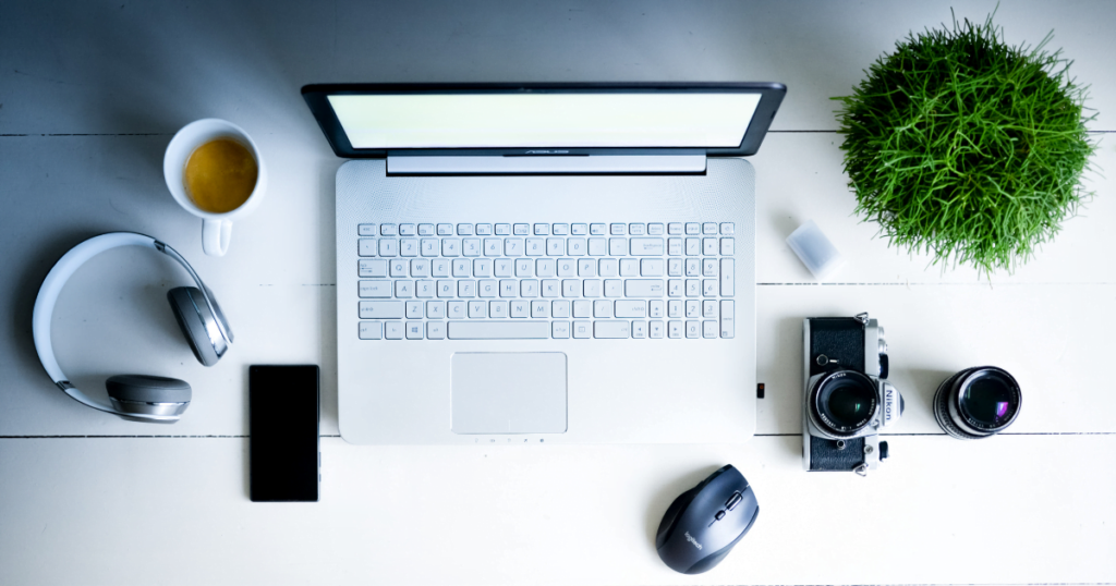 A birds eye view of a white desk with common office supplies, including a laptop, headphones, camera and lens, a mouse, a coffee cup, and a plant.