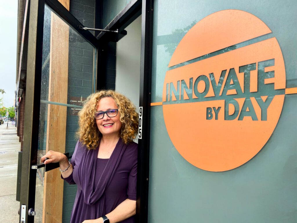 Deb Day smiling in the doorway of the Innovate By Day office, beside the orange Innovate By Day sign