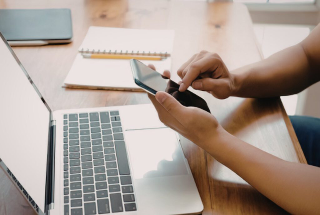 Person scrolling a smartphone beside an open laptop and notebook